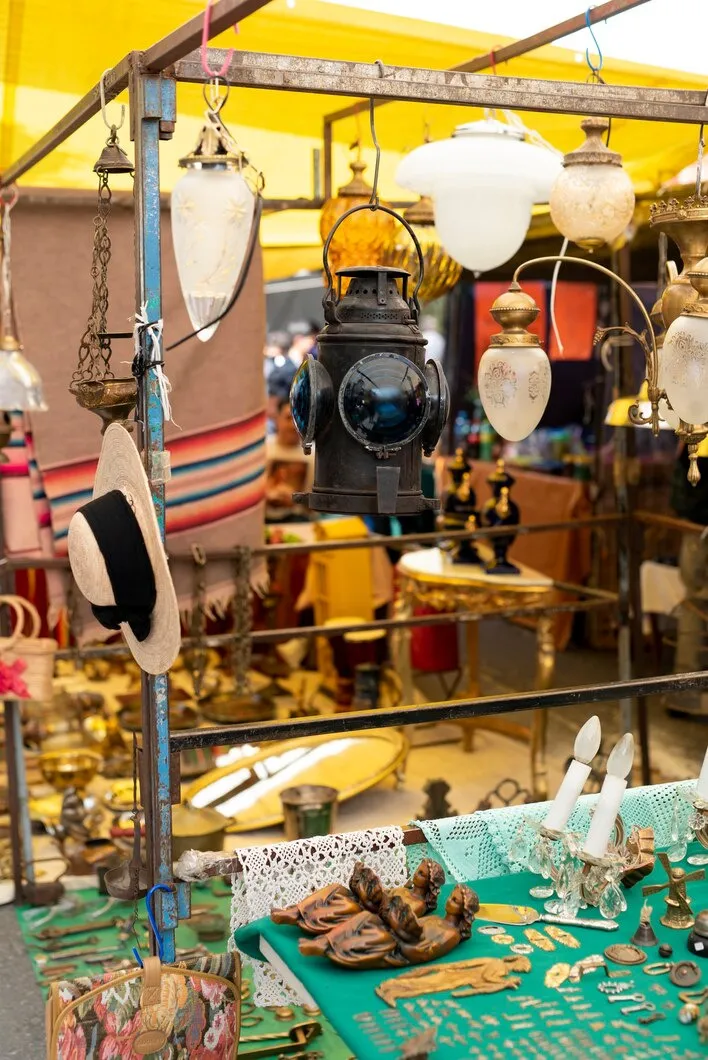 Marché aux puces avec divers objets anciens exposés : lanternes, chapeau, bibelots, bougeoirs et nappes en crochet.
